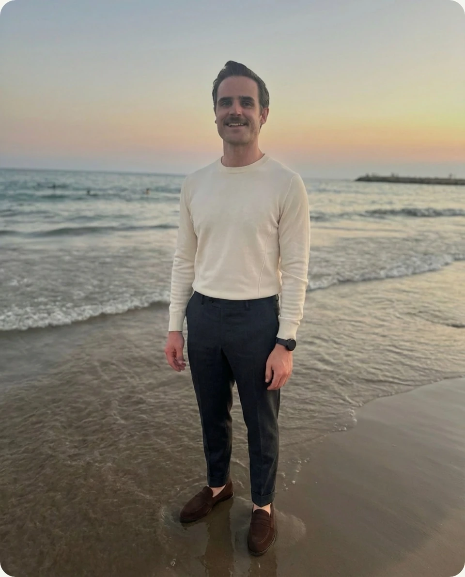 Thraed try-on: a man on a beach at sunset wearing the cream sweater, charcoal trousers and brown loafers